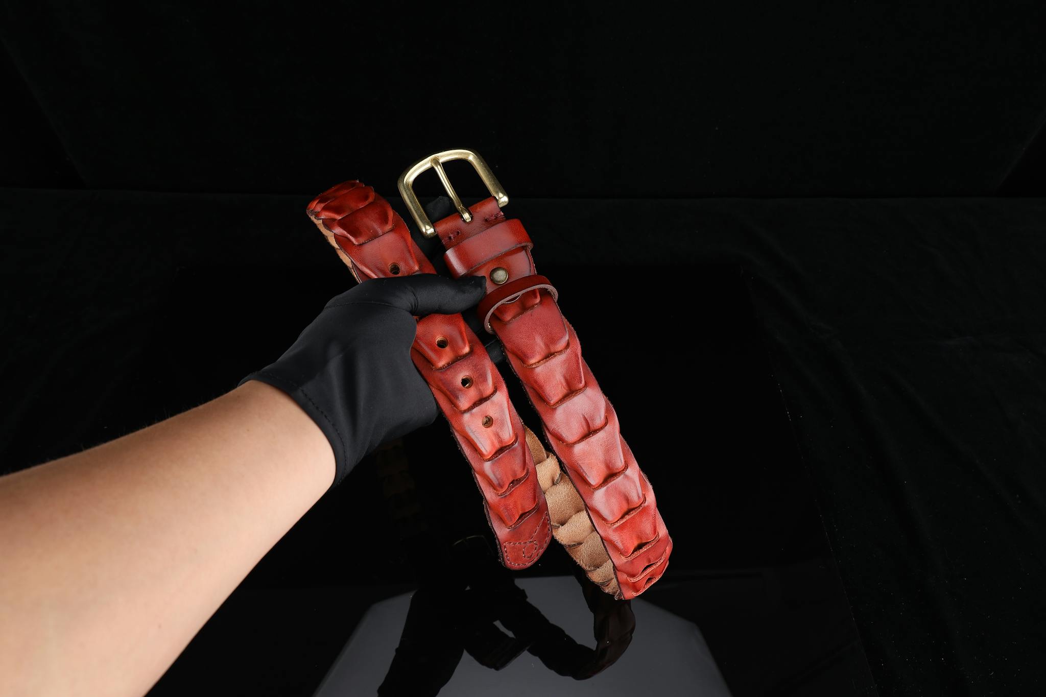 A red leather belt with a gold buckle is elegantly held by a gloved hand against a black background.