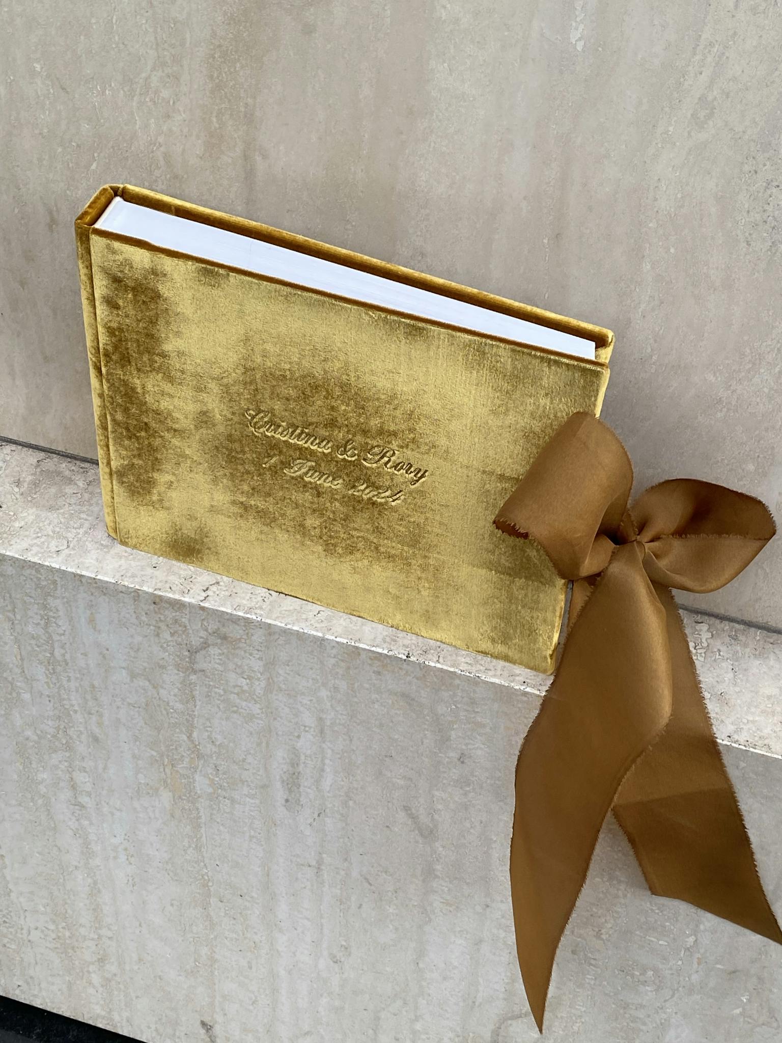 Gold velvet book with a brown silk bow on a marble surface, offering a luxurious gift presentation.